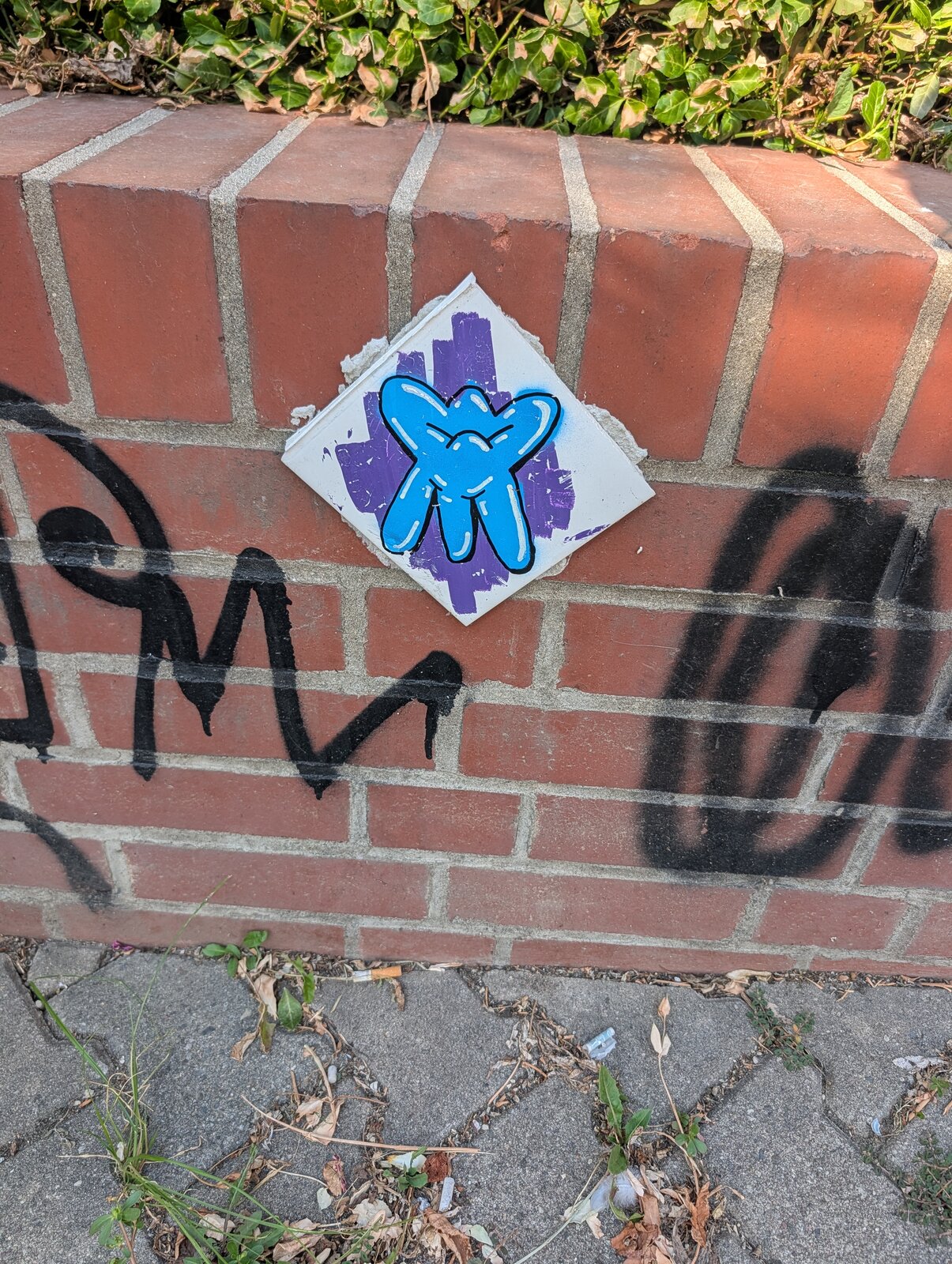 Ein blaues figürliches Motiv auf weißem Papier mit violettem Hintergrund, aufgeklebt auf einer roten Backsteinmauer. Daneben sind schwarze Graffiti-Tags zu sehen.