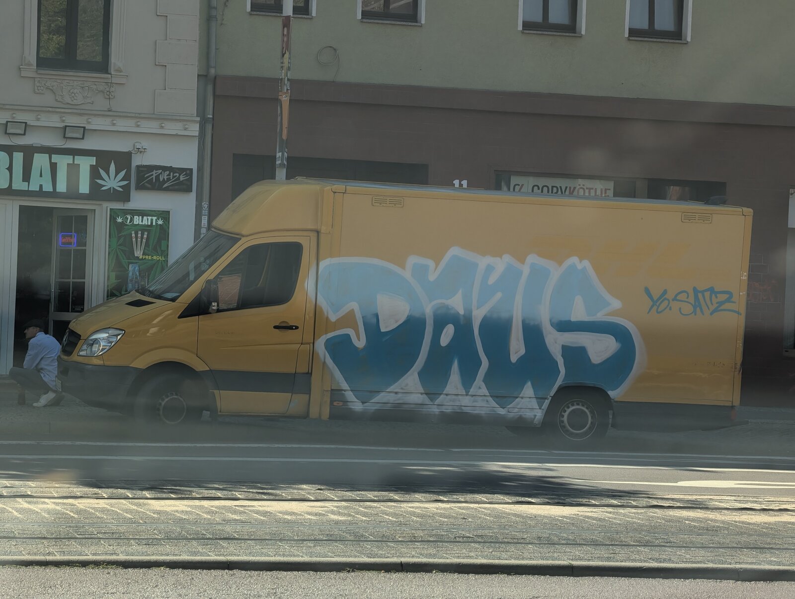 Gelber Transporter mit blauem Graffiti-Schriftzug 'DAVIS' auf der Seite, daneben ein kleineres Tag 'YOSHI'. Im Hintergrund Wohngebäude mit Geschäften, darunter ein Laden mit Cannabisblatt-Logo.
