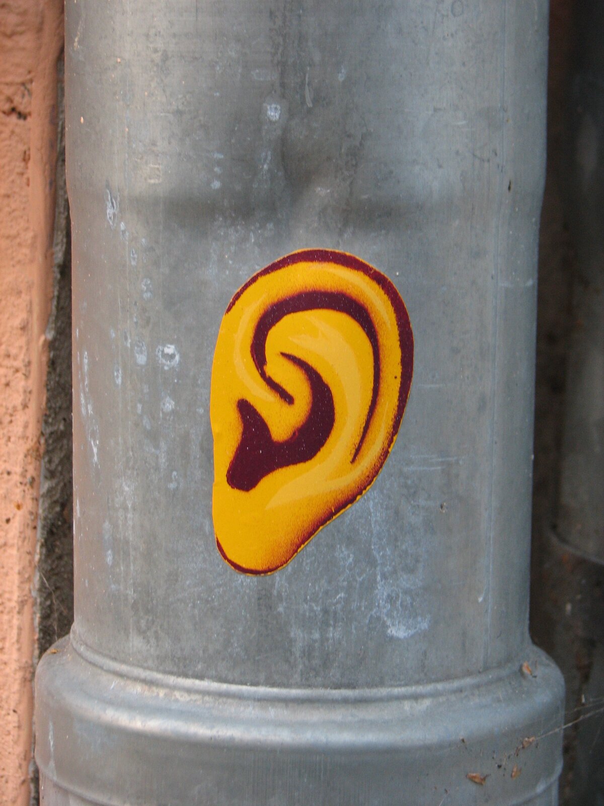 Ein gelbes Ohr mit dunkelroter Kontur auf grauem Untergrund, aufgebracht auf einer zylindrischen Betonsäule neben einer rötlichen Backsteinwand.