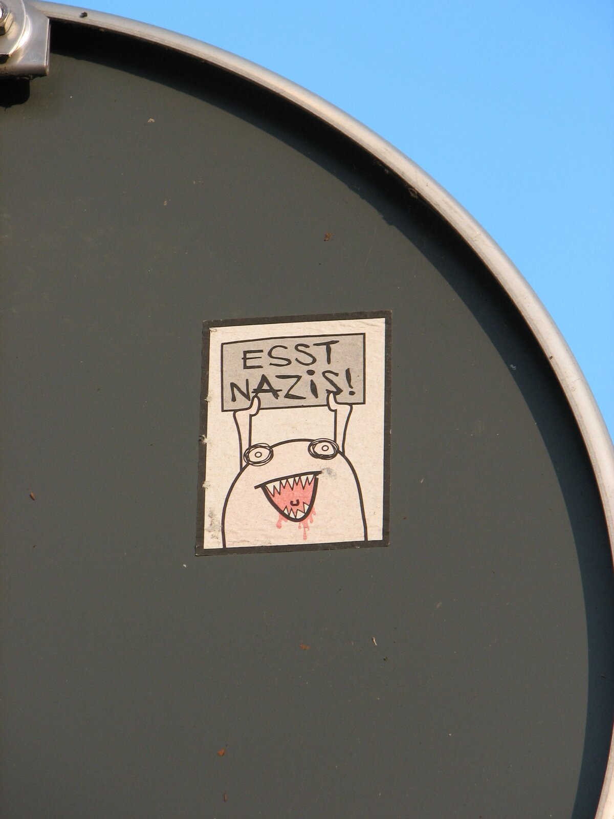 Ein Sticker auf grauer Oberfläche zeigt eine einfache Zeichnung einer Figur mit ovalem Kopf und offenem Mund. Der Text "ESST NAZIS" ist darüber geschrieben. Die Zeichnung ist in Schwarz-Weiß mit rosa Akzenten im Mundbereich.