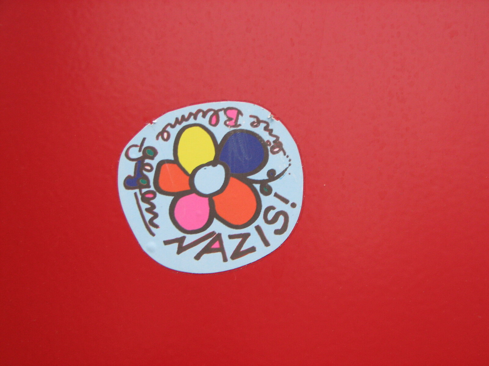 Ein runder Sticker auf rotem Untergrund mit einer bunten Blüte in der Mitte und handschriftlichem Text (eine Blume gegen Nazis) im Kreis herum in Braun und Dunkelblau.