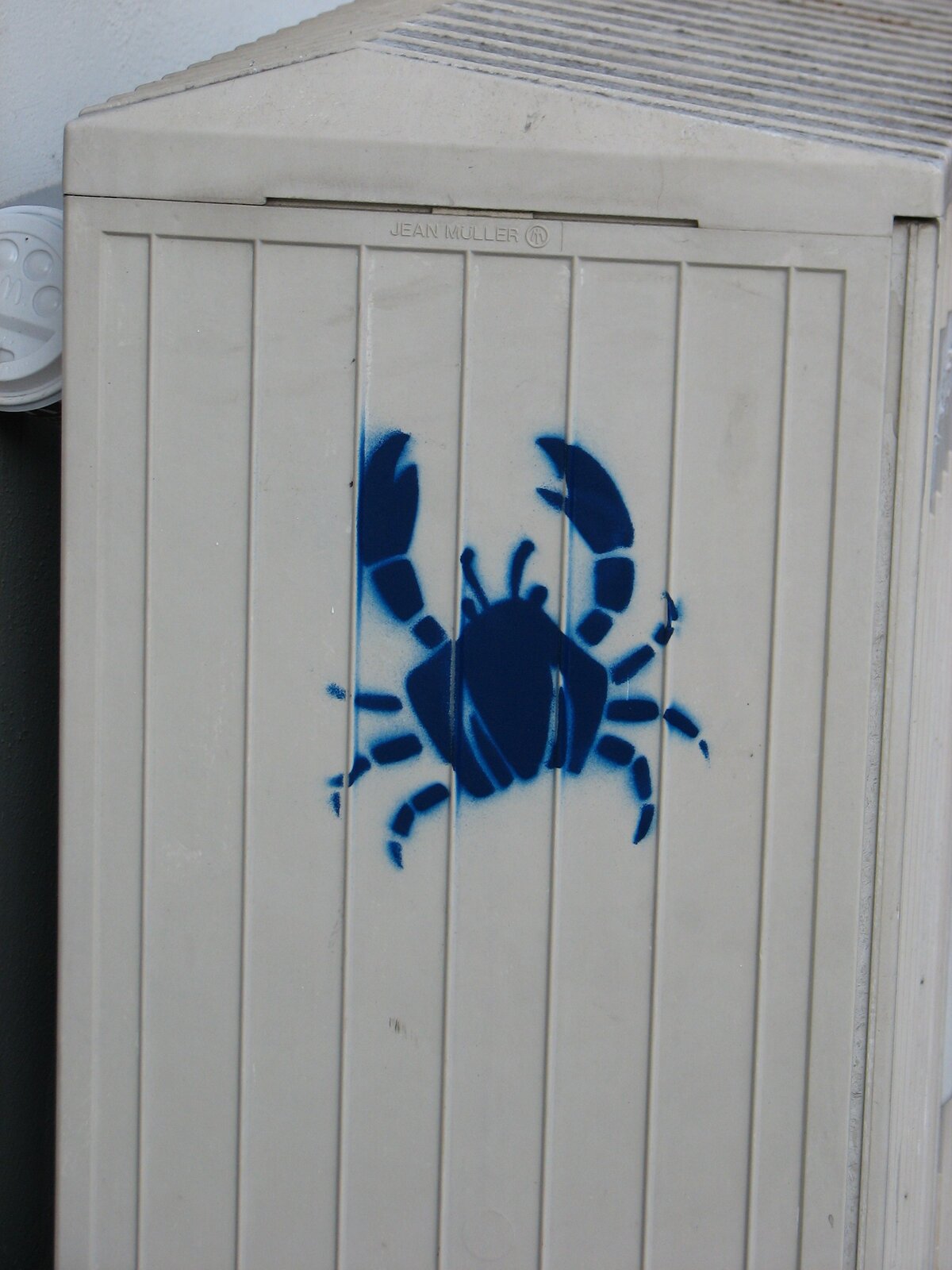 Blausprühte Stencil-Figur eines Krebses auf weißer Oberfläche. Das Motiv zeigt das Tier in Draufsicht mit ausgebreiteten Scheren und Beinen, gesprüht auf vertikalen Metallplatten.