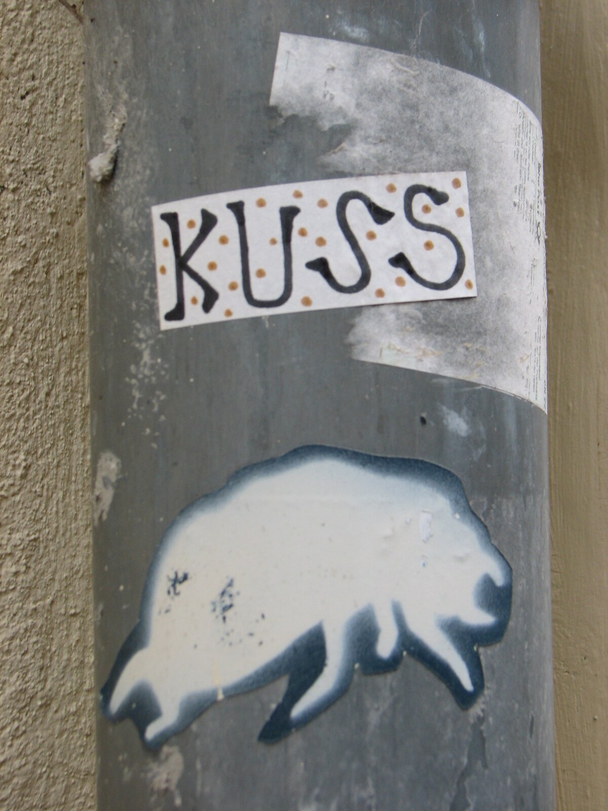 Weißer rechteckiger Sticker mit schwarzer Schrift 'KUSS' und dekorativen Punkten, darunter ein schwarzes Stencil-Motiv eines Insekts.