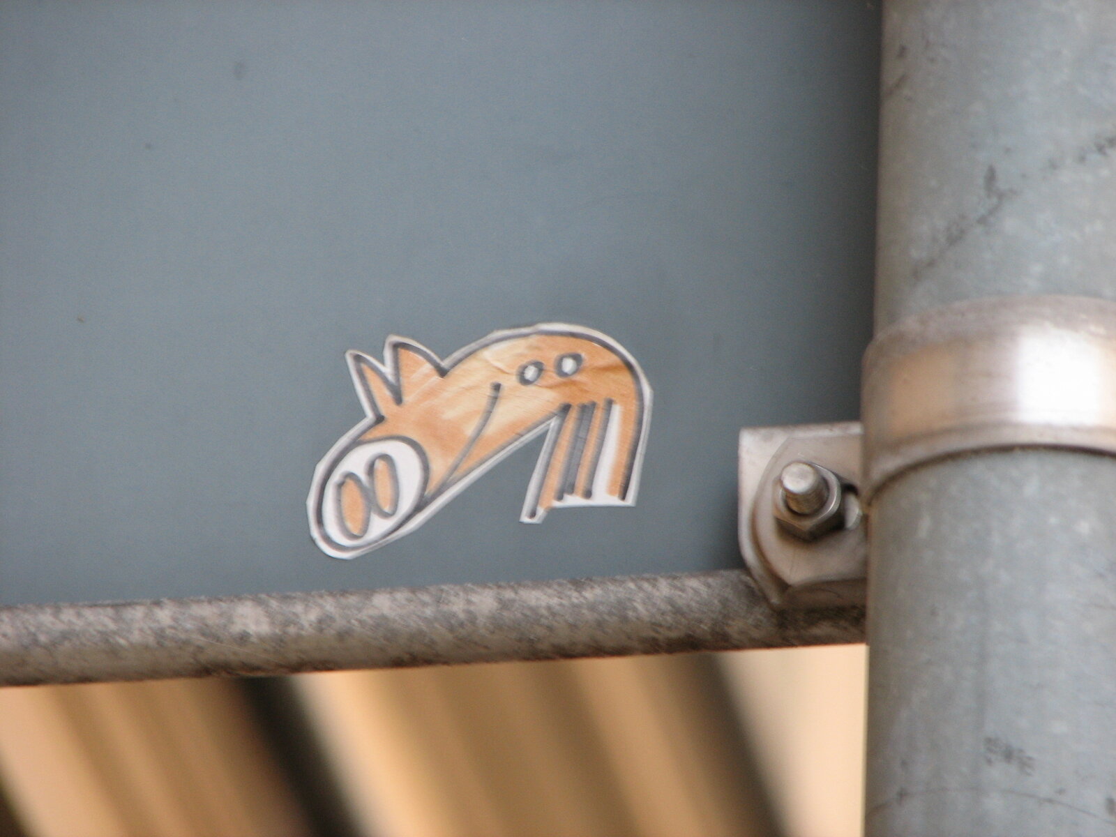 Orangefarbener Sticker auf grauer Metallfläche mit stilisiertem Tiermotiv und weißer Kontur, angebracht an einer Rohrkonstruktion.